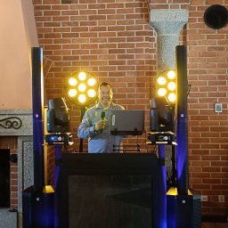 Dj.Marciz - Uśmiechnięty mężczyzna za konsoletą DJ-ską z laptopem i mikrofonem, otoczony kolumnami głośnikowymi i reflektorami na tle ceglanej ściany.