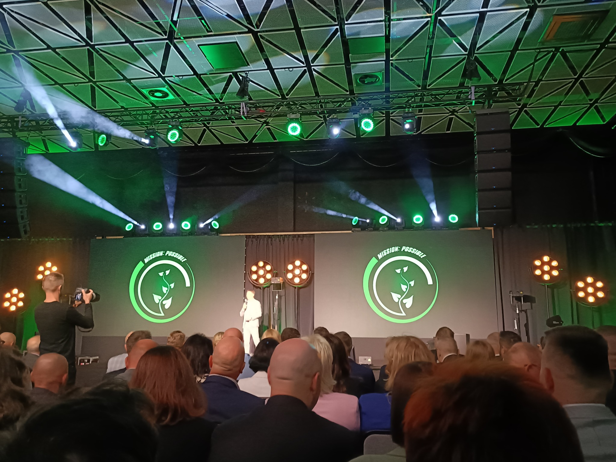 Konferencja z prelegentem na scenie oświetlonej zielonymi światłami i reflektorami, widoczny tłum słuchaczy oraz operator kamery.
