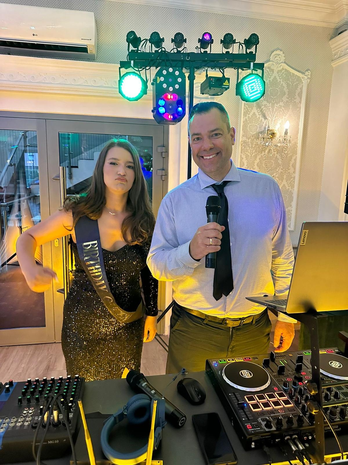 DJ z mikrofonem i dziewczyna z szarfą urodzinową pozują na tle sprzętu nagłośnieniowego i oświetlenia imprezowego.