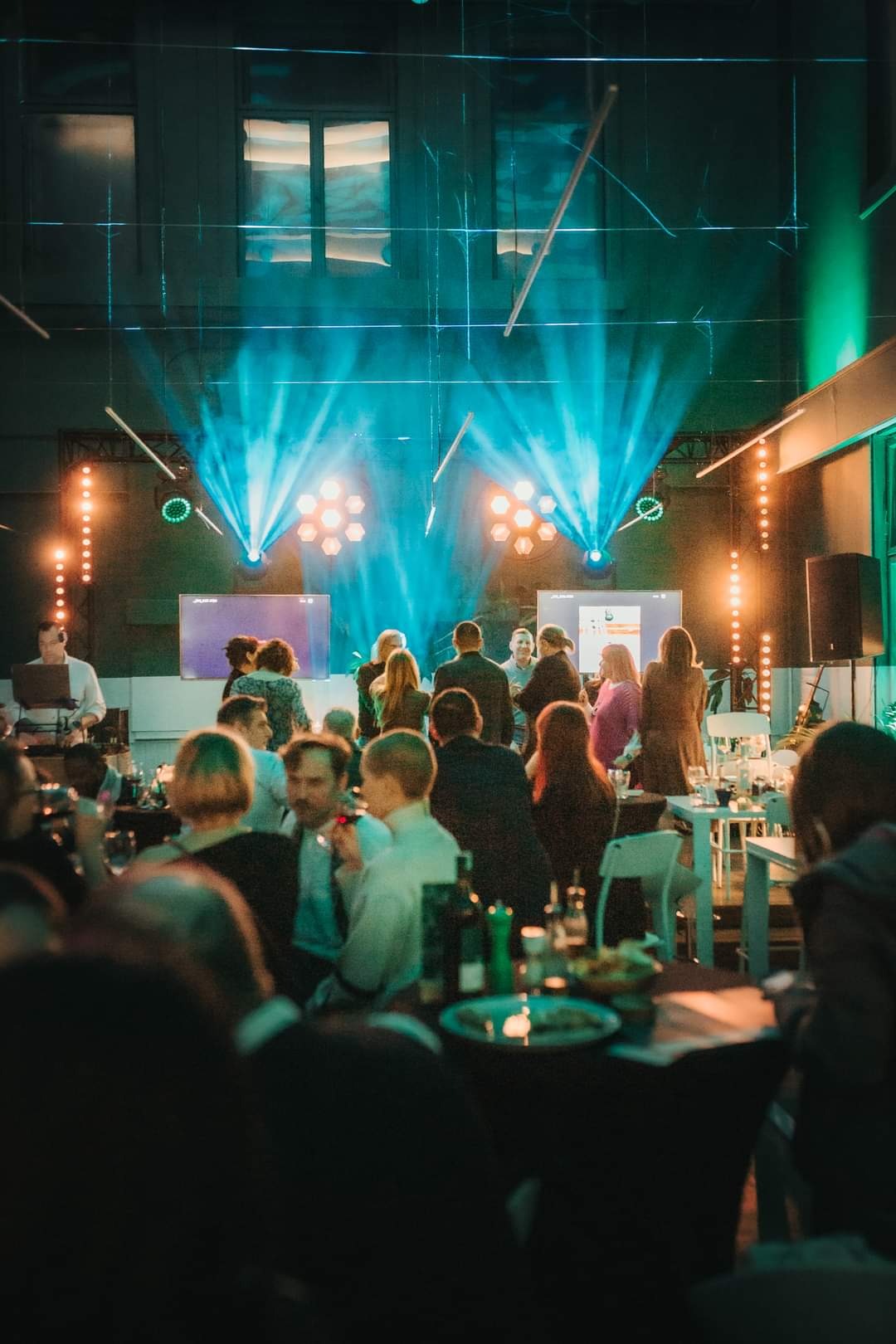 Wnętrze sali konferencyjnej podczas wydarzenia z oświetleniem scenicznym, widoczne stoły z cateringiem i publiczność.