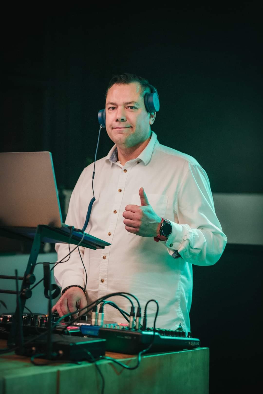 Mężczyzna w słuchawkach daje kciuk w górę, stoi za konsoletą DJ-ską z laptopem, oświetlony zielonym światłem.