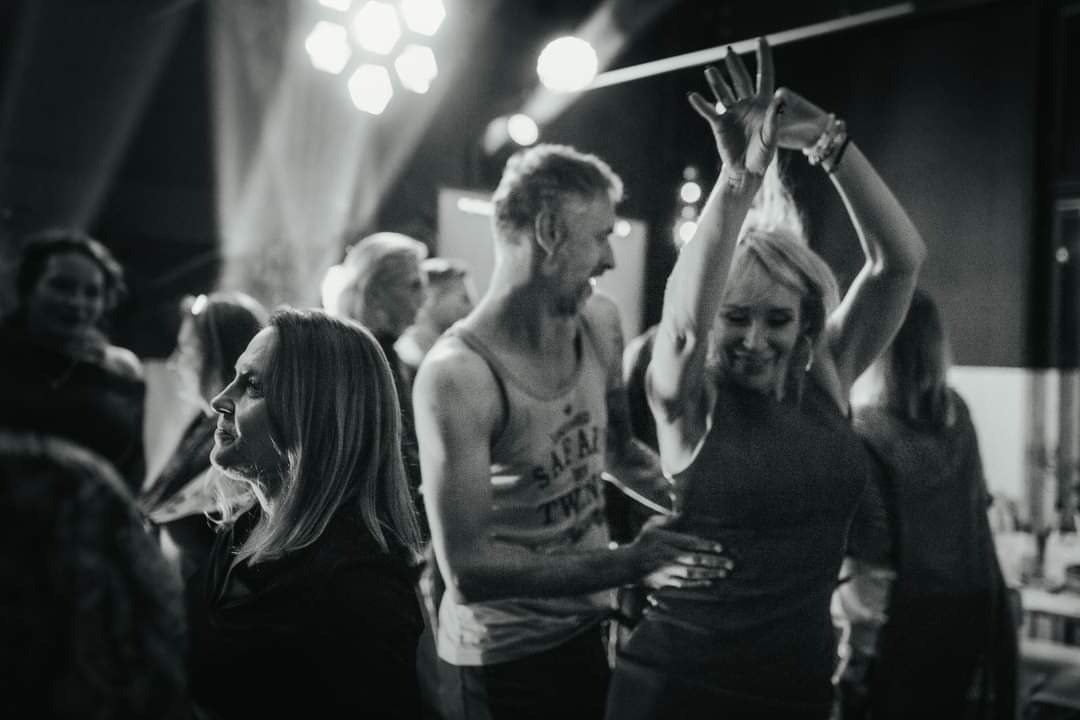 Czarno-białe zdjęcie przedstawia rozbawionych gości na imprezie, tańczących w blasku reflektorów. W tle widoczne osoby, a na pierwszym planie kobieta z podniesionymi rękami i uśmiechem na twarzy.