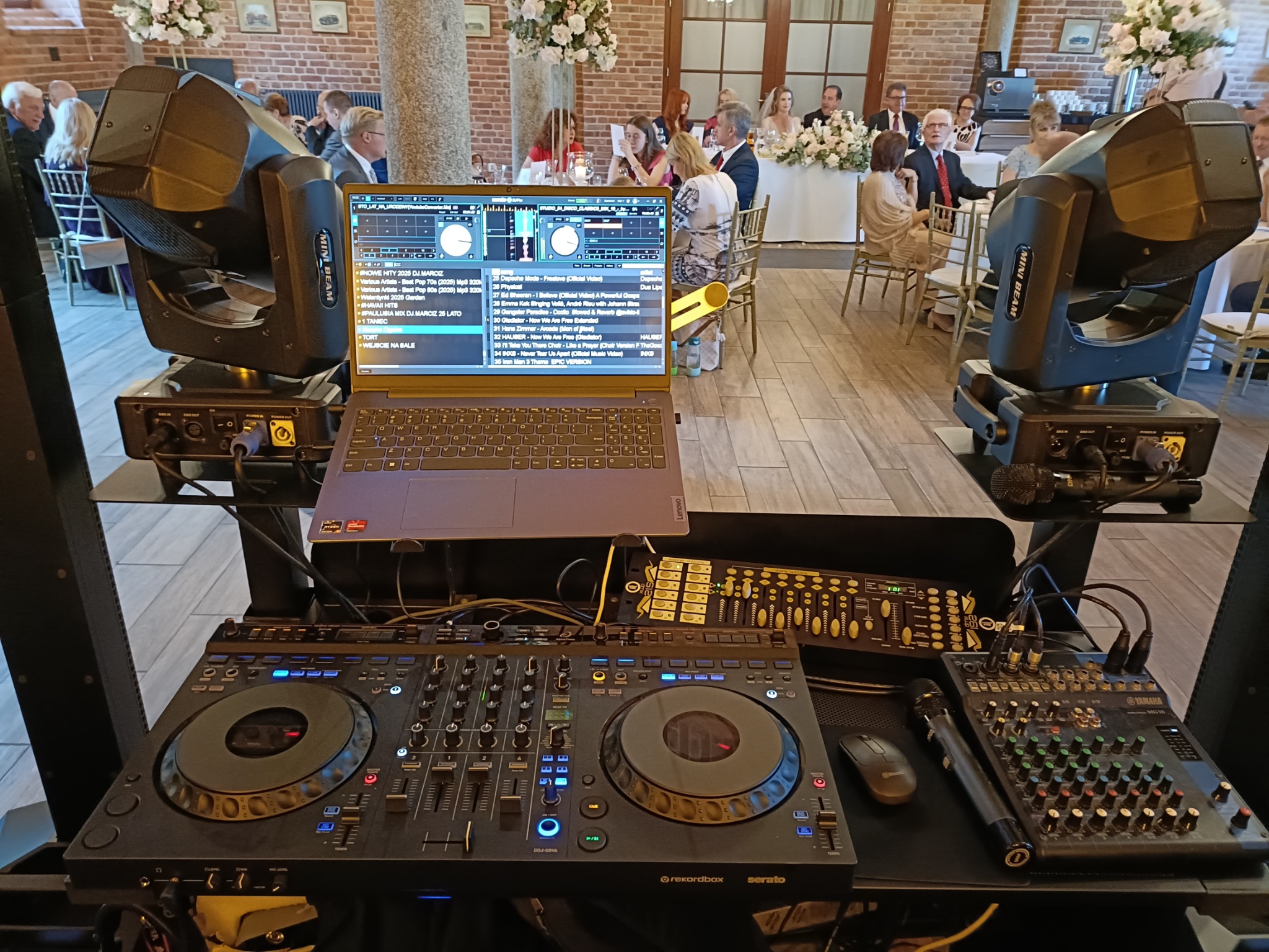 DJ stanowisko z laptopem, mikserem i oświetleniem na sali weselnej. Widoczni goście w tle. Sprzęt gotowy do występu, oświetlenie ustawione.