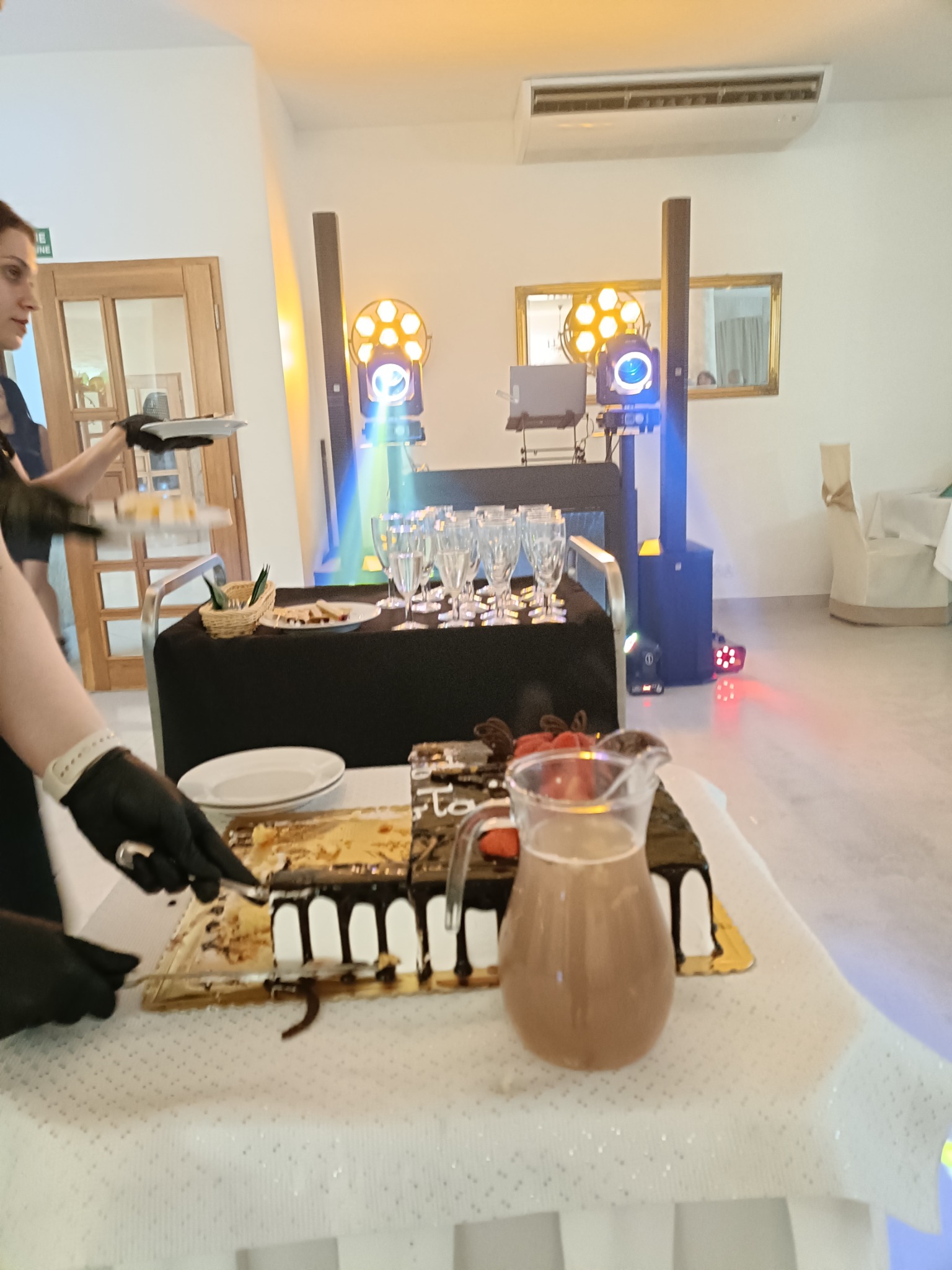 Krojenie tortu na przyjęciu z nagłośnieniem i oświetleniem. Kelnerka w czarnych rękawiczkach serwuje tort. W tle profesjonalne oświetlenie eventowe i szklanki.