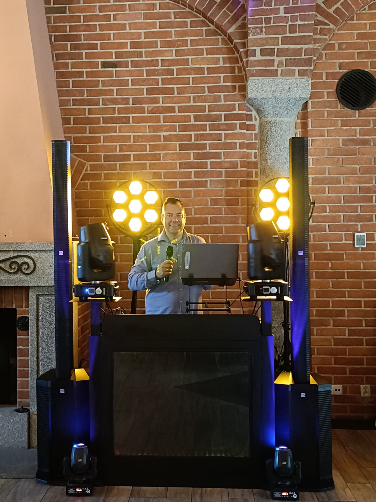Uśmiechnięty mężczyzna za konsoletą DJ-ską z laptopem i mikrofonem, otoczony kolumnami głośnikowymi i reflektorami na tle ceglanej ściany.