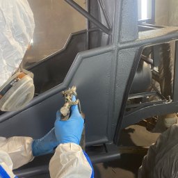 Car-Czyścik Daniel Ziaja - Malowanie natryskowe ramy pojazdu w kolorze szarym, pracownik w kombinezonie ochronnym i niebieskich rękawicach, używa pistoletu lakierniczego.