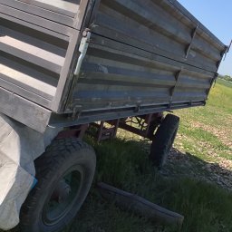 Car-Czyścik Daniel Ziaja - Przyczepa rolnicza z metalową skrzynią ładunkową, widoczna z boku na tle zielonej trawy i błękitnego nieba, z elementami konstrukcyjnymi i oponami w średnim stanie technicznym.