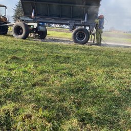 Car-Czyścik Daniel Ziaja - Operator w kombinezonie ochronnym czyści przyczepę rolniczą z użyciem strumienia ściernego na tle zielonej łąki.