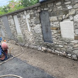 Car-Czyścik Daniel Ziaja - Sprzęt do czyszczenia strumieniowo-ściernego (piaskowania) stoi przed kamiennym murem z wmurowanymi płytami nagrobnymi, widoczne ślady czyszczenia muru.