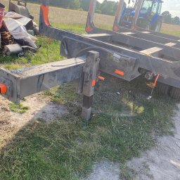 Car-Czyścik Daniel Ziaja - Przyczepa leśna z niebieskim traktorem w tle, widoczne elementy konstrukcyjne przyczepy, przewody elektryczne i opony, na trawiastym podłożu.