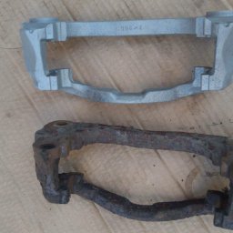 Car-Czyścik Daniel Ziaja - Porównanie: Stary, zardzewiały zacisk hamulcowy obok nowego, pomalowanego na srebrno zacisku; widoczne ślady korozji na starym elemencie.