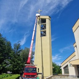 Car-Czyścik Daniel Ziaja - Czerwony podnośnik koszowy na tle kościelnej wieży z krzyżem, błękitne niebo z delikatnymi chmurami, widok z dołu na budynek kościoła i chodnik z kostki.