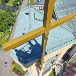 Car-Czyścik Daniel Ziaja - Widok z góry na złoty krzyż na dachu kościoła, cień robotników na krzyżu, samochód z agregatem na placu, zielone pola w tle.