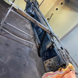 Car-Czyścik Daniel Ziaja - Widok z dołu na fragment maszyny przemysłowej z metalowymi rurami i elementami hydraulicznymi, częściowo osłoniętej kolorową tkaniną, obok metalowy regał.