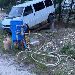 Car-Czyścik Daniel Ziaja - Niebieski mobilny agregat do obróbki strumieniowo-ściernej z podłączonymi przewodami, na tle białego busa ustawionego na podwyższeniu i worków z materiałem ściernym.