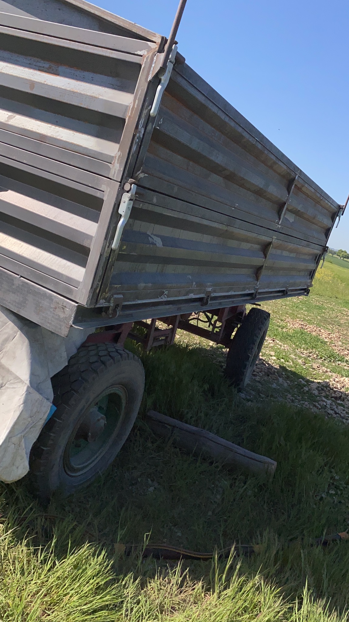 Przyczepa rolnicza z metalową skrzynią ładunkową, widoczna z boku na tle zielonej trawy i błękitnego nieba, z elementami konstrukcyjnymi i oponami w średnim stanie technicznym.