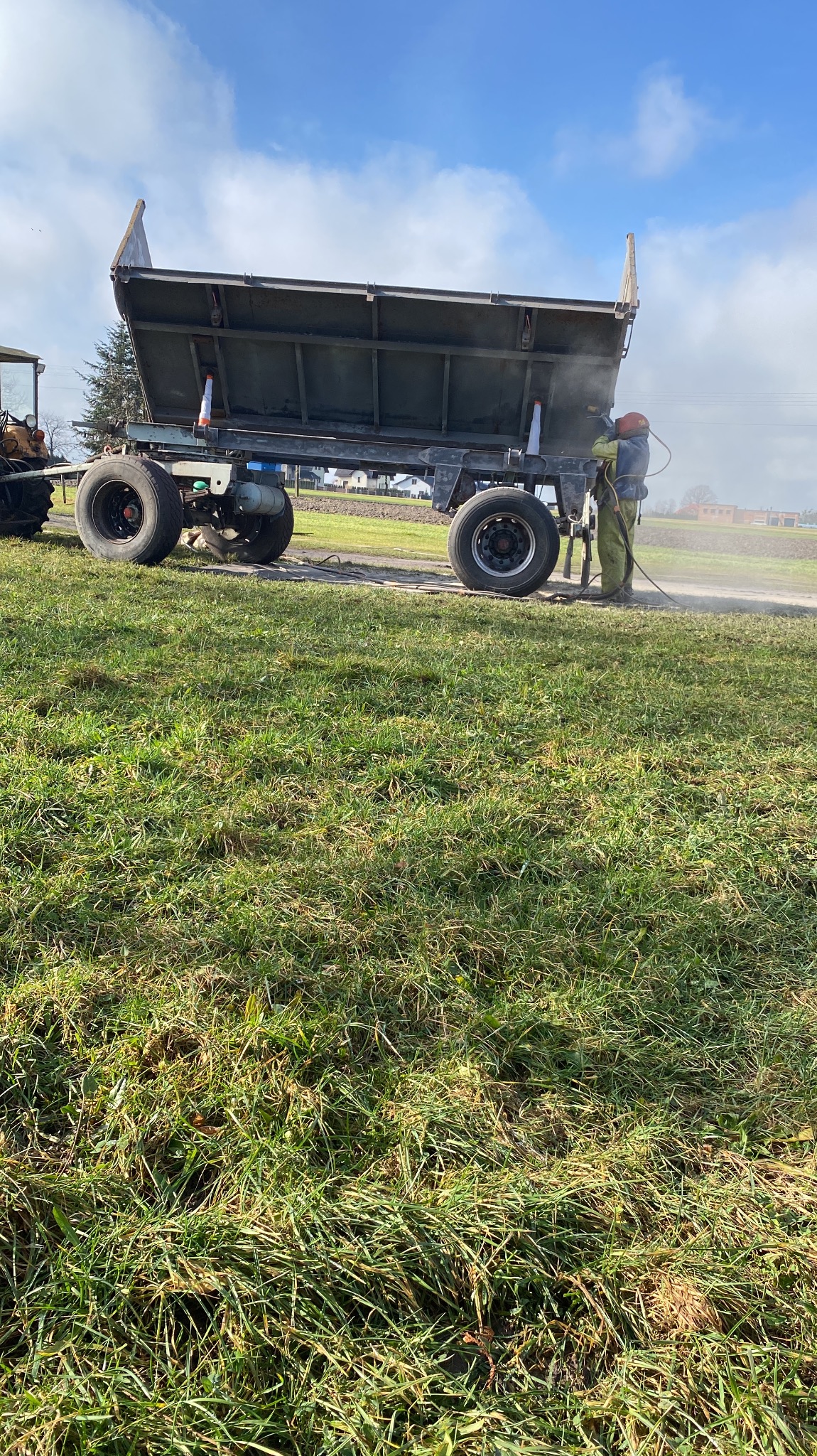 Operator w kombinezonie ochronnym czyści przyczepę rolniczą z użyciem strumienia ściernego na tle zielonej łąki.