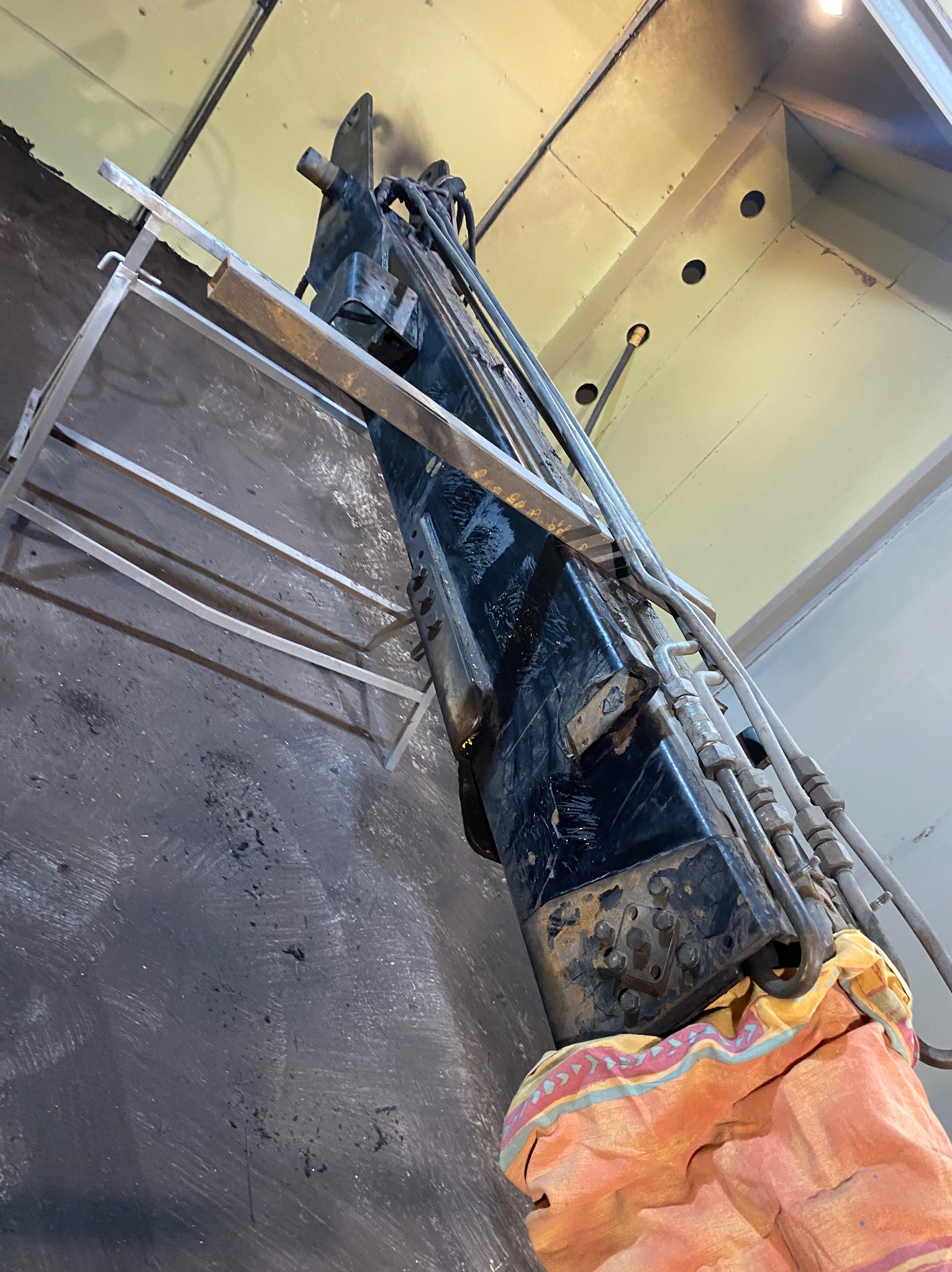 Czarny, przemysłowy sprzęt z rurami hydraulicznymi, częściowo owinięty kolorowym materiałem, stoi obok metalowego rusztowania w pomieszczeniu o jasnym suficie z widocznymi otworami.