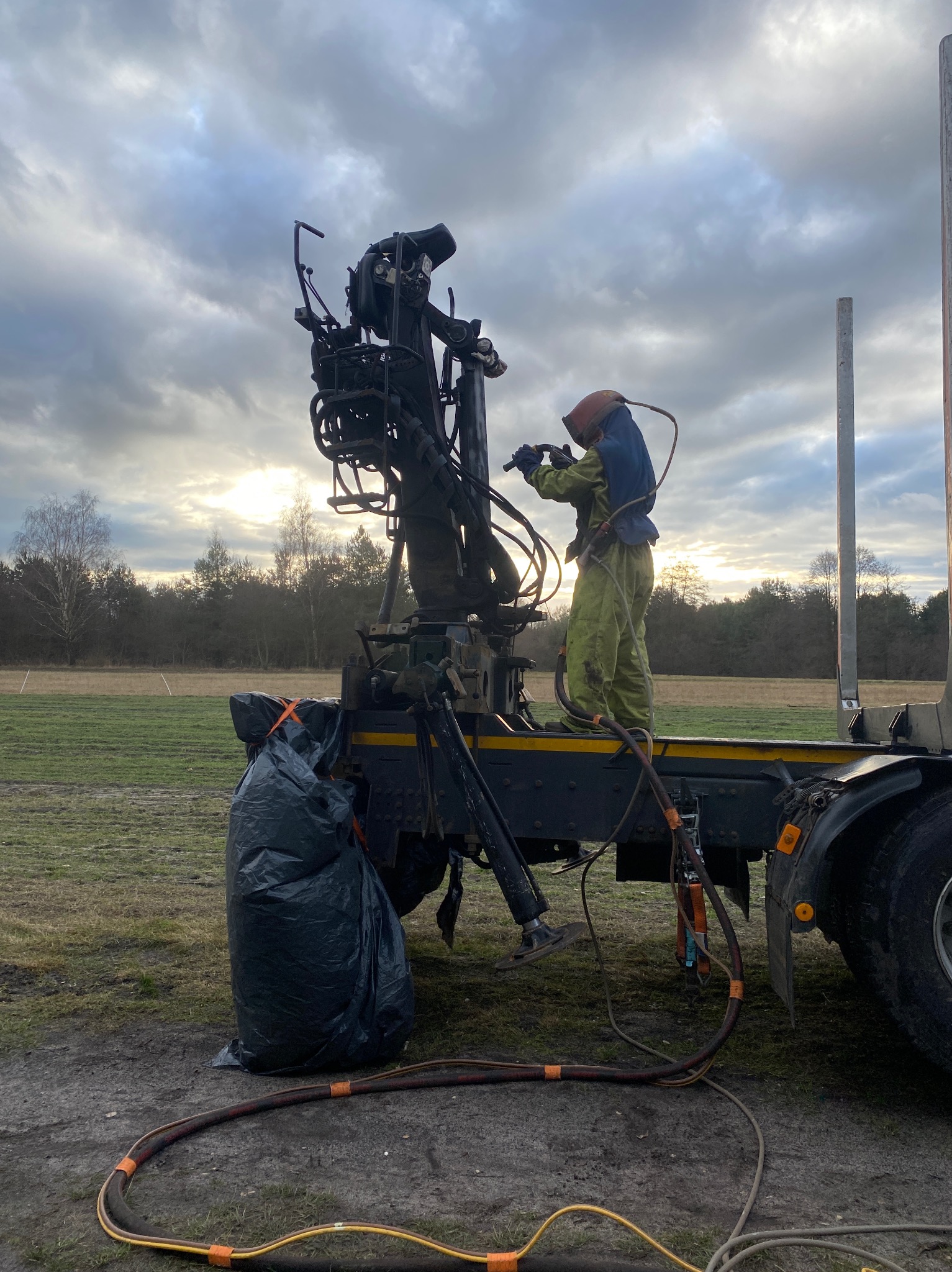 Pracownik w kombinezonie ochronnym i hełmie piaskuje konstrukcję na platformie ciężarówki; w tle łąka i las pod pochmurnym niebem.