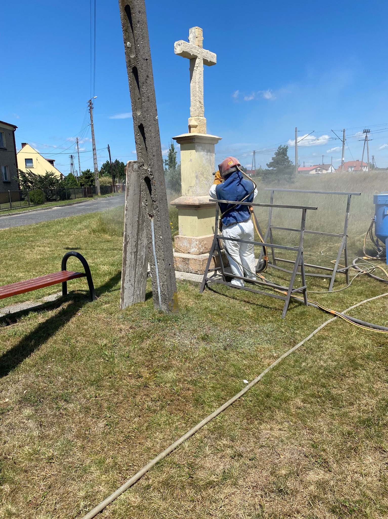 Pracownik w kombinezonie ochronnym i hełmie oczyszcza krzyż przydrożny za pomocą strumienia ściernego, obok metalowa konstrukcja i wąż. W tle zabudowania wiejskie.