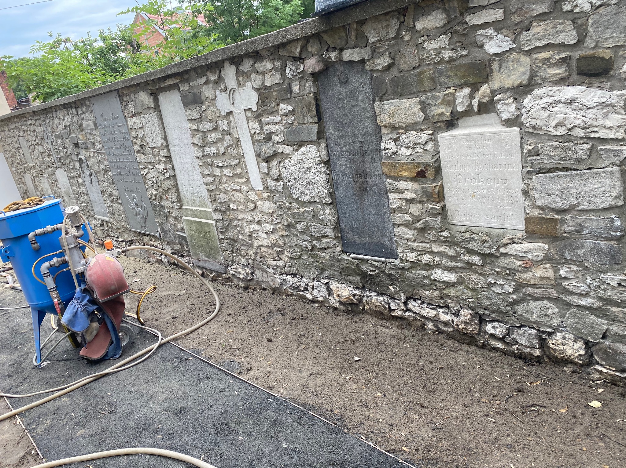 Sprzęt do czyszczenia strumieniowo-ściernego (piaskowania) stoi przed kamiennym murem z wmurowanymi płytami nagrobnymi, widoczne ślady czyszczenia muru.