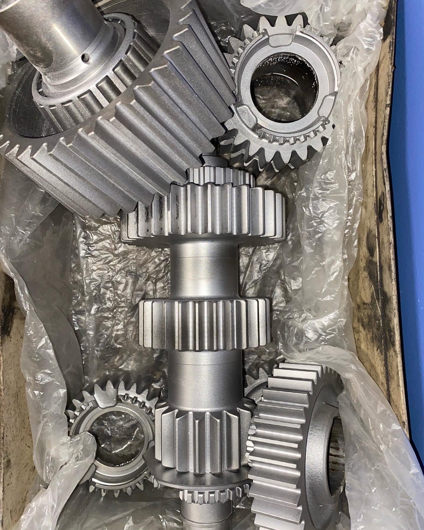 Metalowe koła zębate, w tym jedno z dwoma rzędami zębów na wale, po obróbce, leżące w pudełku wyłożonym folią.