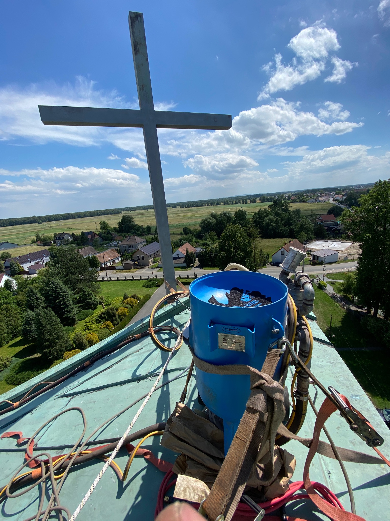 Piaskowanie krzyża na dachu kościoła, widoczny niebieski zbiornik piaskarki, kable i liny zabezpieczające, w tle panorama miejscowości i błękitne niebo z chmurami.