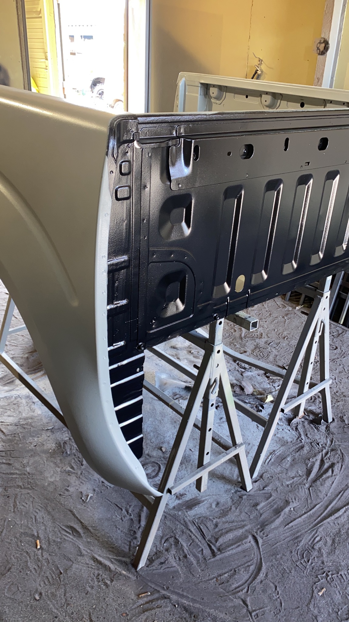 Element karoserii samochodu typu pickup, po nałożeniu czarnej powłoki antykorozyjnej, umieszczony na metalowych kozłach w warsztacie.