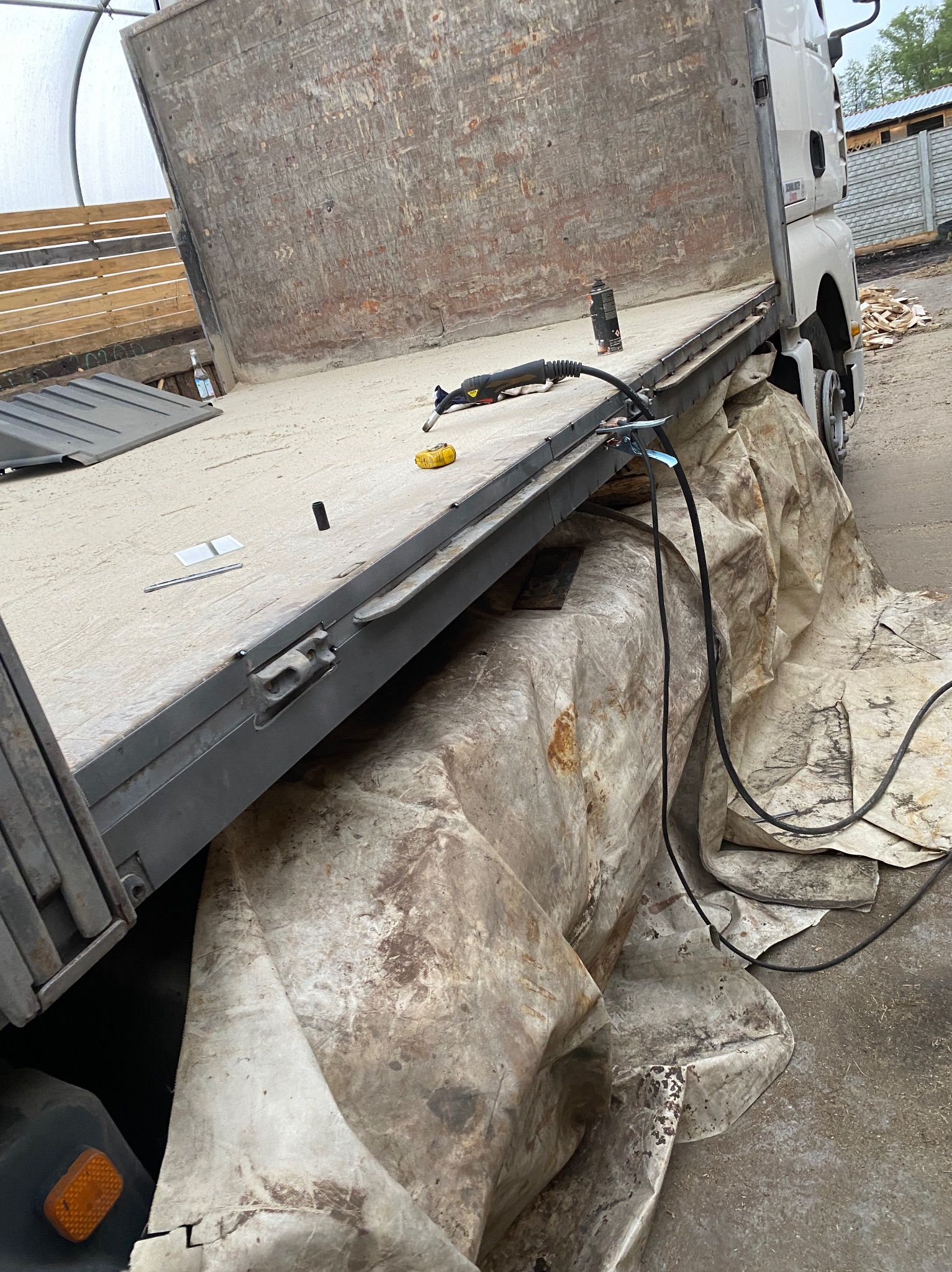 Platforma samochodu ciężarowego z widocznymi śladami spawania i narzędziami spawalniczymi, zabezpieczona plandeką, w tle budynek gospodarczy i stos drewna.