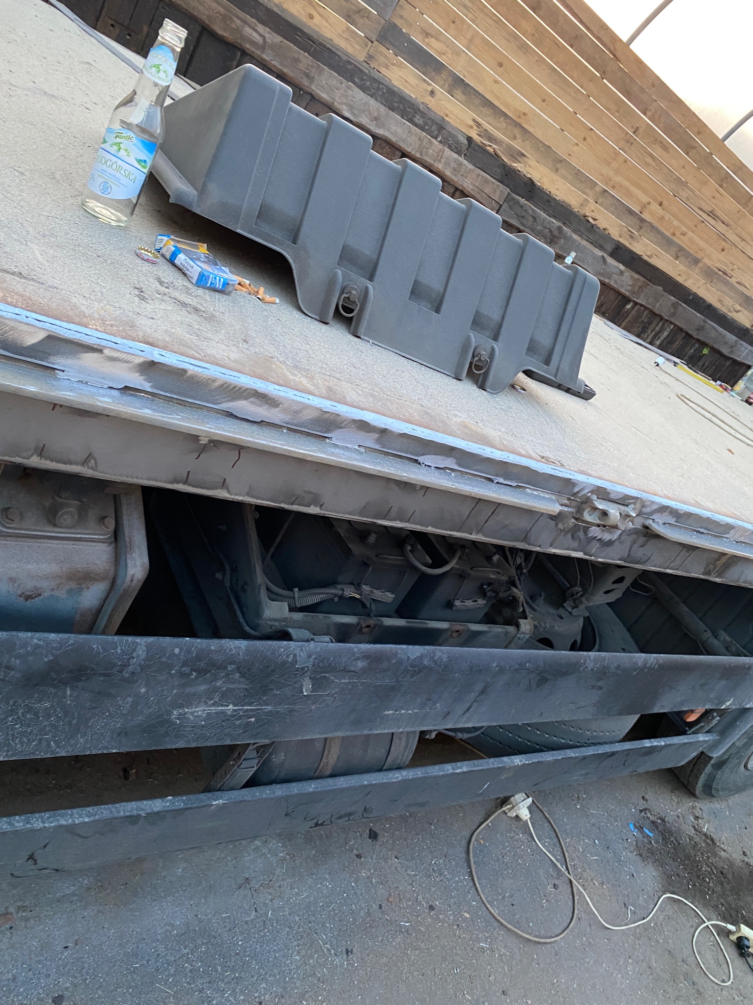 Fragment podwozia samochodu ciężarowego z widocznymi elementami zawieszenia, elektrycznym kablem zasilającym i plastikową skrzynką. Na podłodze leżą niedopałki papierosów oraz butelka po napoju.