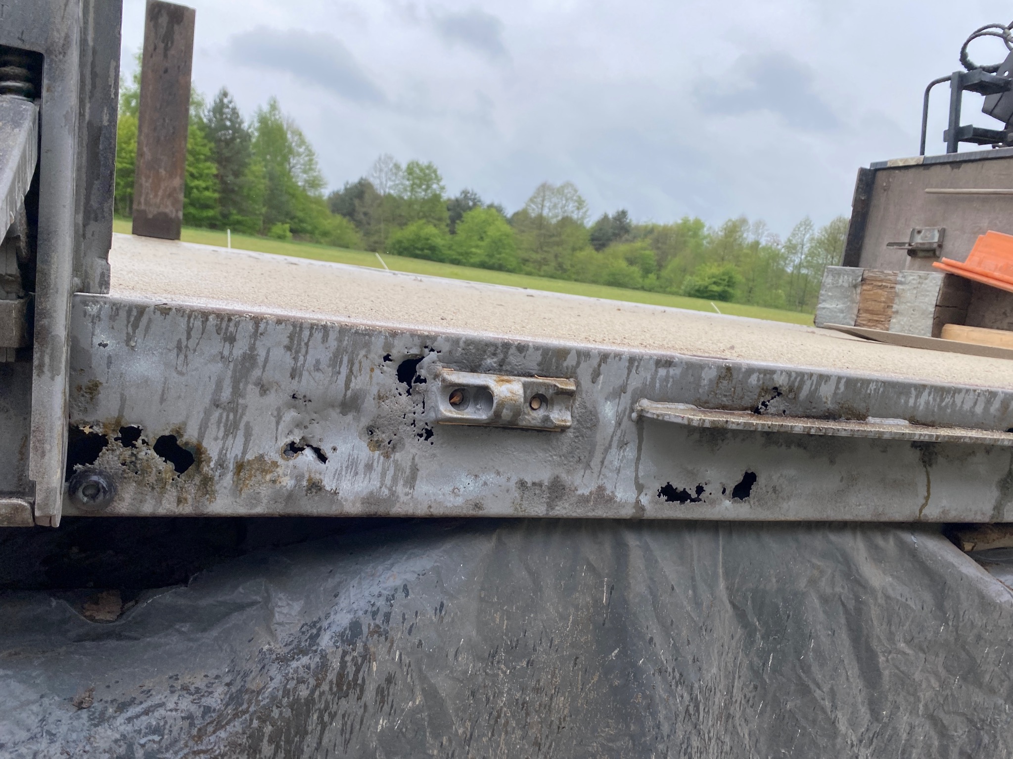 Fragment skorodowanej, metalowej burty samochodu ciężarowego z widocznymi dziurami i śladami rdzy, na tle zieleni i pochmurnego nieba.