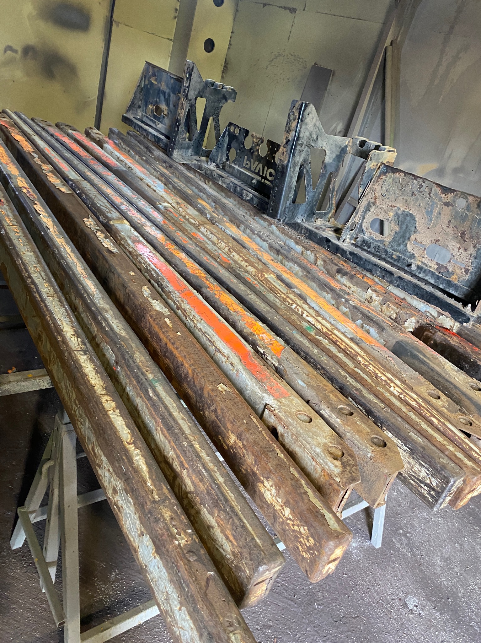 Rzędy zardzewiałych, metalowych belek o przekroju kwadratowym, niektóre z resztkami pomarańczowej farby, ułożone na metalowym stojaku w warsztacie, czekające na piaskowanie lub konserwację...