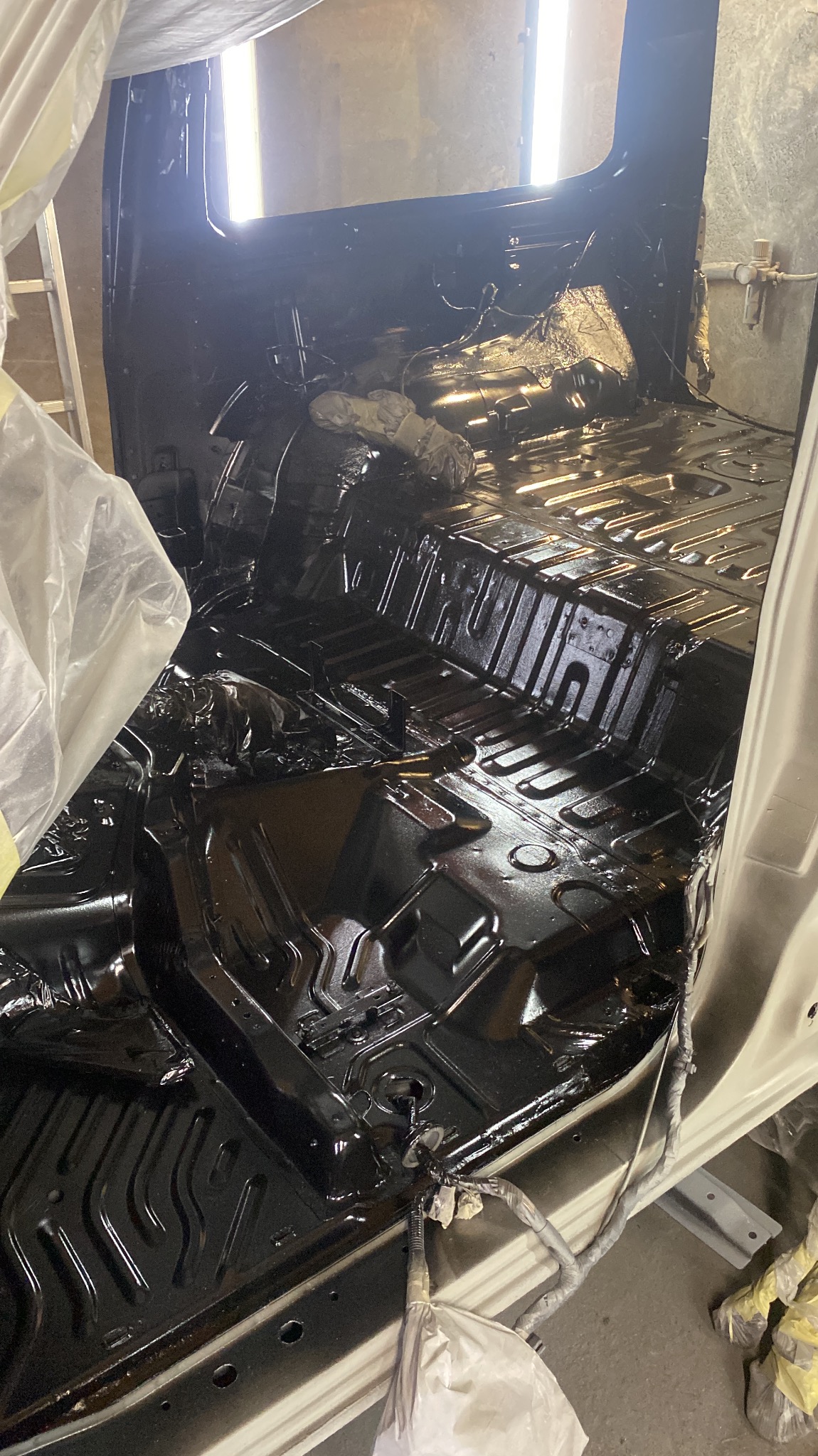 Wnętrze samochodu po nałożeniu czarnej powłoki ochronnej, widoczne ślady świeżego lakieru, zabezpieczone elementy karoserii folią i taśmą malarską.