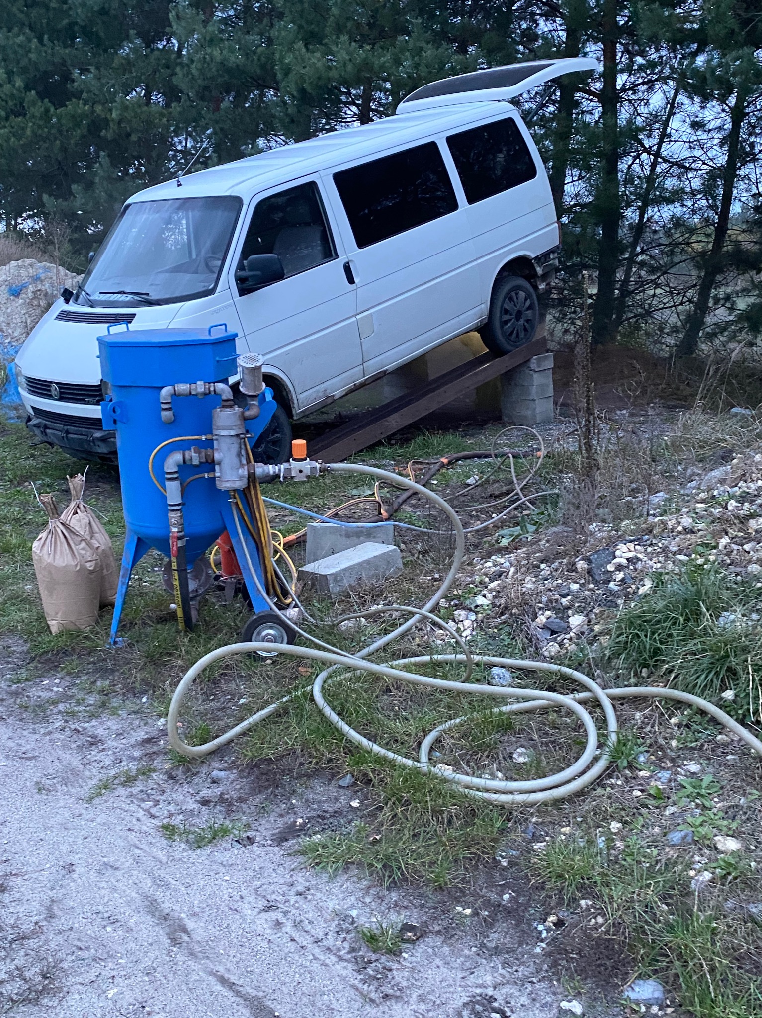 Niebieski mobilny agregat do obróbki strumieniowo-ściernej z podłączonymi przewodami, na tle białego busa ustawionego na podwyższeniu i worków z materiałem ściernym.