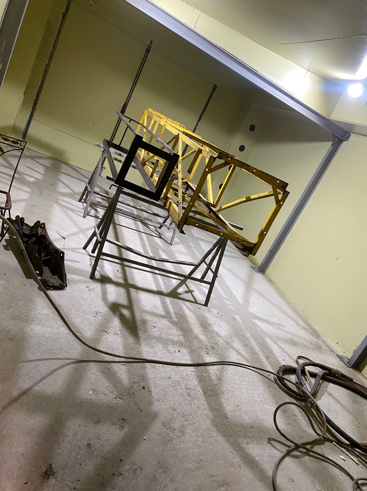Wnętrze hali z żółtą metalową konstrukcją na kozłach, czarnym elementem ramy i przewodami na podłodze, prawdopodobnie przygotowywanej do obróbki lub malowania.