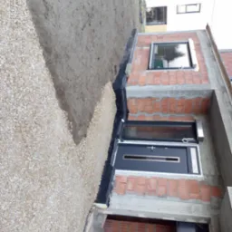 Budynek w trakcie budowy, widoczne fundamenty, ściany z czerwonej cegły i okna, obok wysypany żwir, zabezpieczony czarną folią izolacyjną.