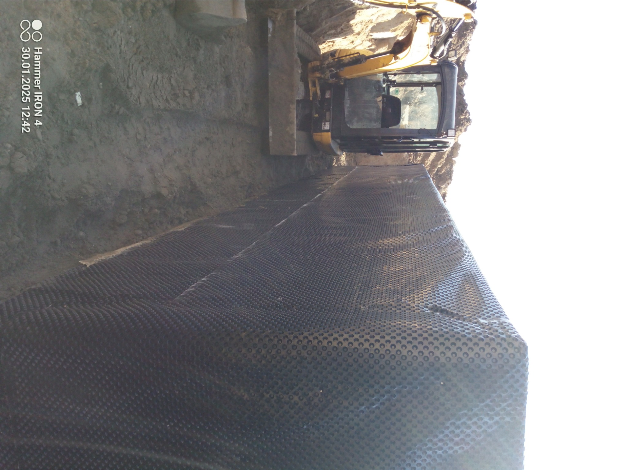 Żółta koparka w wykopie, z widoczną warstwą folii kubełkowej na ścianie wykopu.