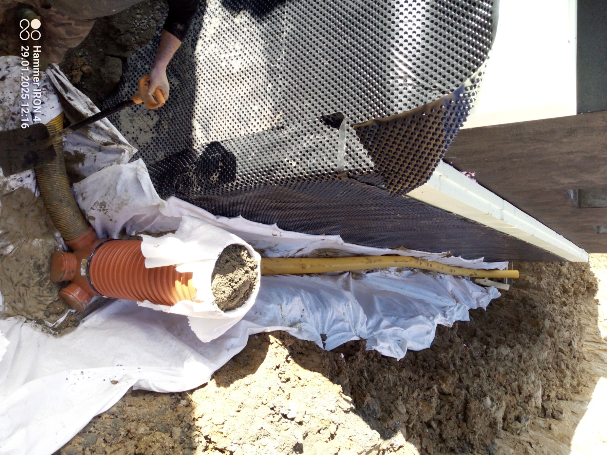 Instalacja rury drenarskiej w wykopie, zabezpieczona geowłókniną i folią kubełkową przy fundamencie budynku, widoczna łopata i warstwa ziemi.