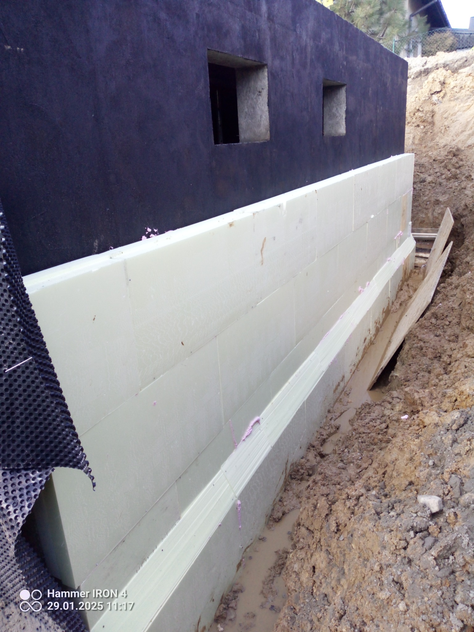 Izolacja termiczna fundamentów z czarną hydroizolacją, widoczny wykop z błotnistą wodą i fragment membrany kubełkowej.