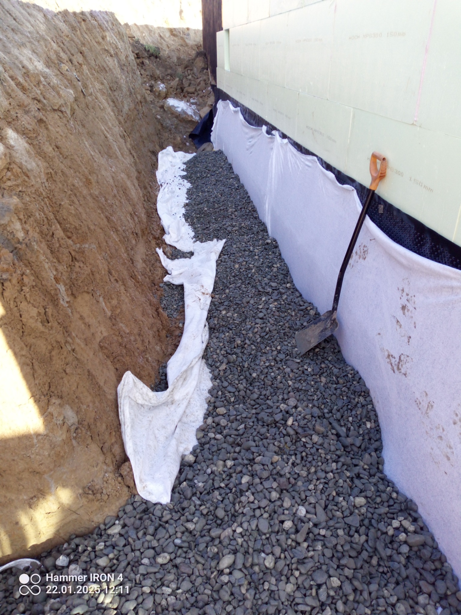 Drenaż fundamentów: wykop z ziemi, warstwa żwiru oddzielona geowłókniną od gruntu, izolacja pionowa fundamentu z polistyrenu ekstrudowanego, widoczna łopata.