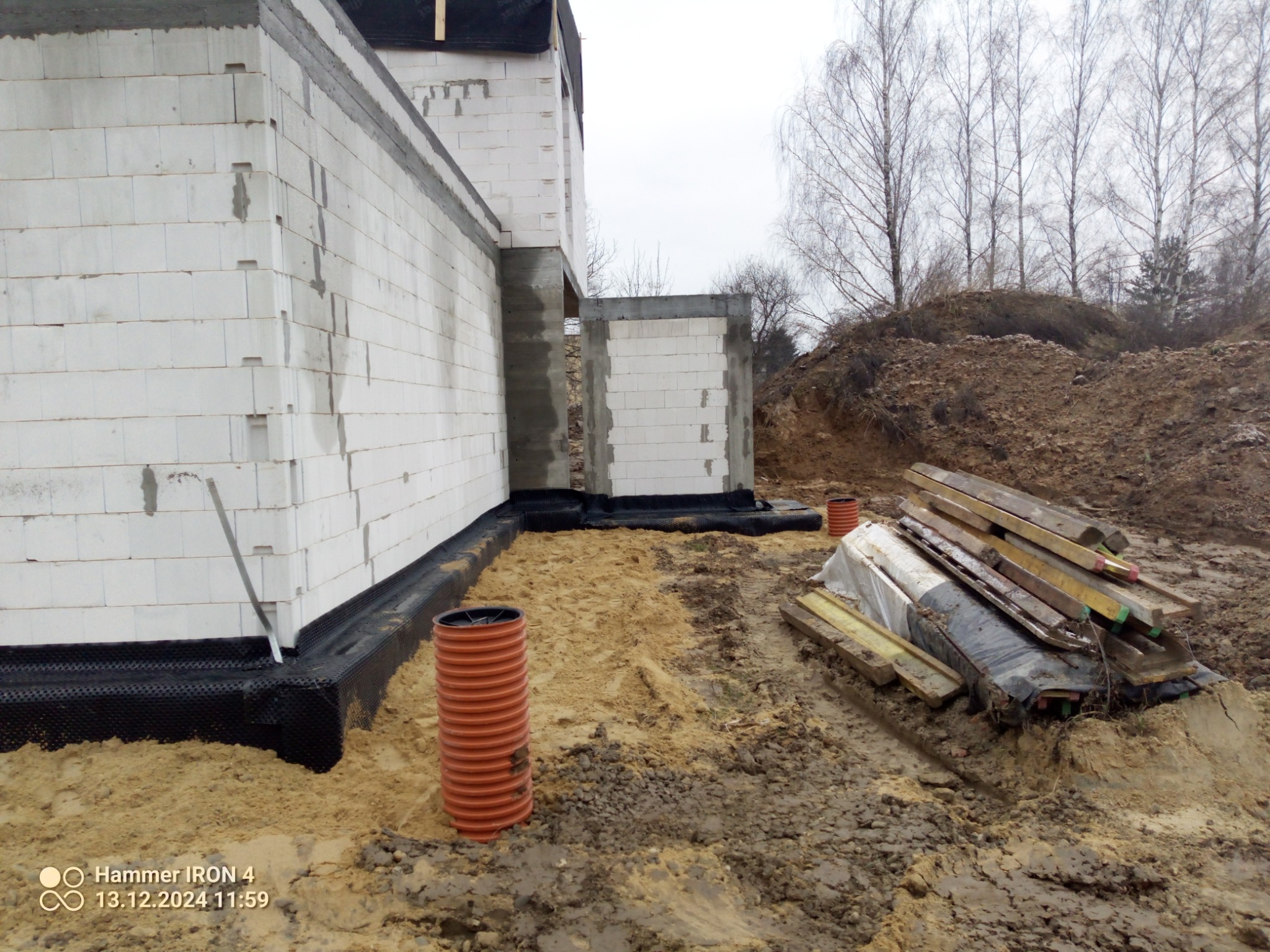 Wznoszone ściany parterowego budynku z białych bloczków, zabezpieczone czarną folią hydroizolacyjną, z widoczną rurą drenarską i stertą drewnianych elementów w surowym otoczeniu placu budowy.