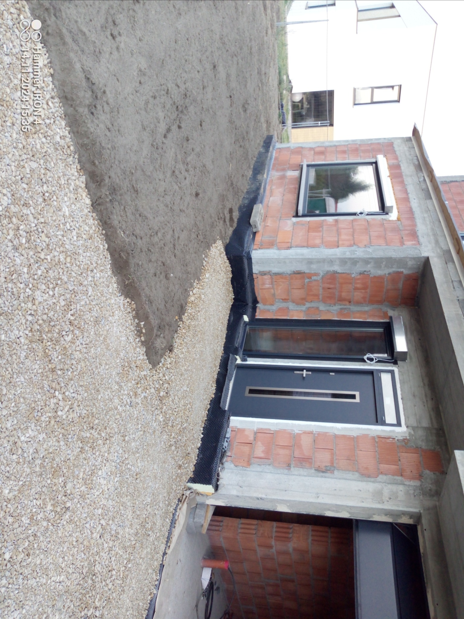 Budynek w trakcie budowy, widoczne fundamenty, ściany z czerwonej cegły i okna, obok wysypany żwir, zabezpieczony czarną folią izolacyjną.