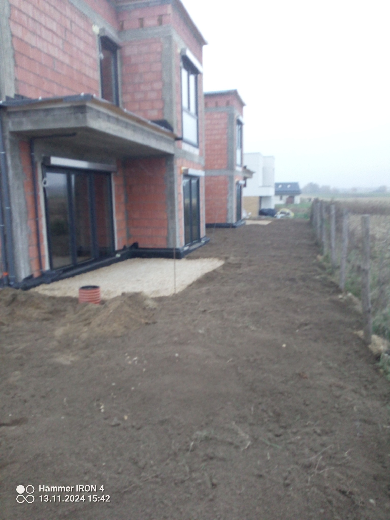 Teren wokół budynków w stanie surowym otwartym, wyrównana ziemia przygotowana pod przyszły ogród, widoczne fragmenty instalacji drenażowej przy fundamentach.