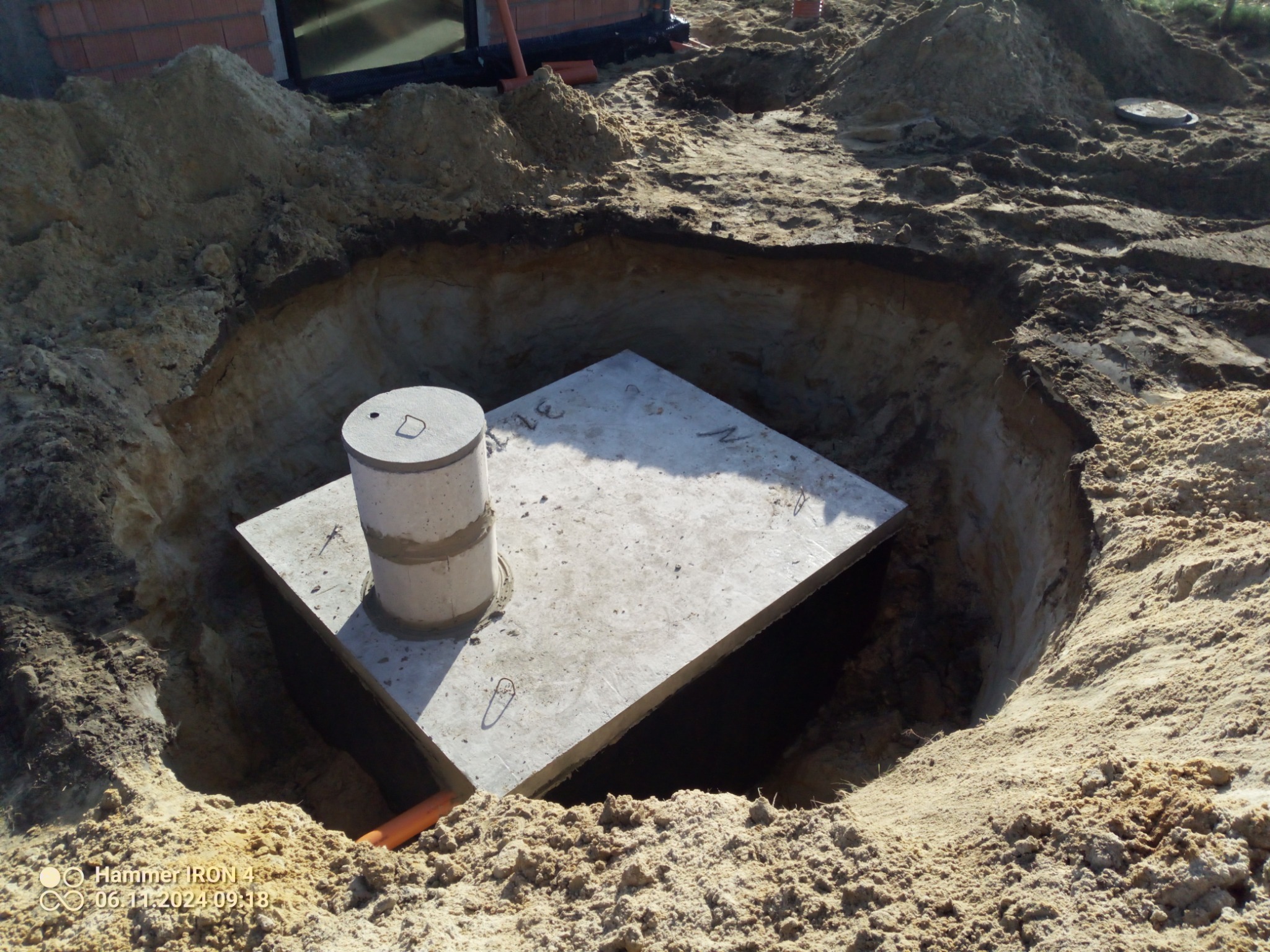 Kwadratowa betonowa płyta z wystającym cylindrycznym elementem w głębokim wykopie ziemnym, widoczny fragment pomarańczowej rury, ziemia usypana w pryzmy wokół wykopu.