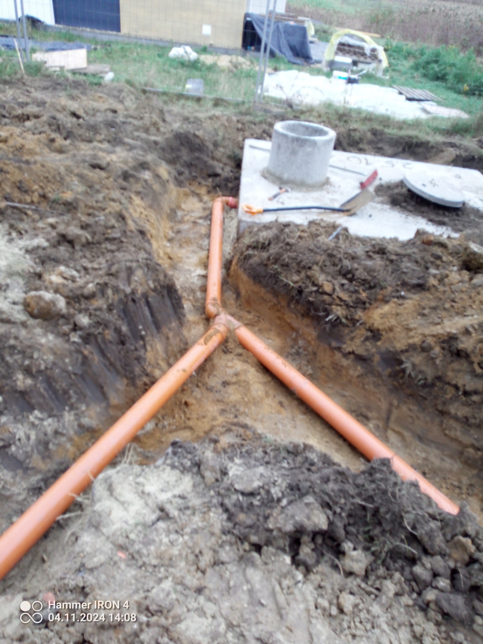 Wykonane rowy w ziemi z pomarańczowymi rurami kanalizacyjnymi układającymi się w kształt litery Y, studzienka betonowa na górze i narzędzia obok.