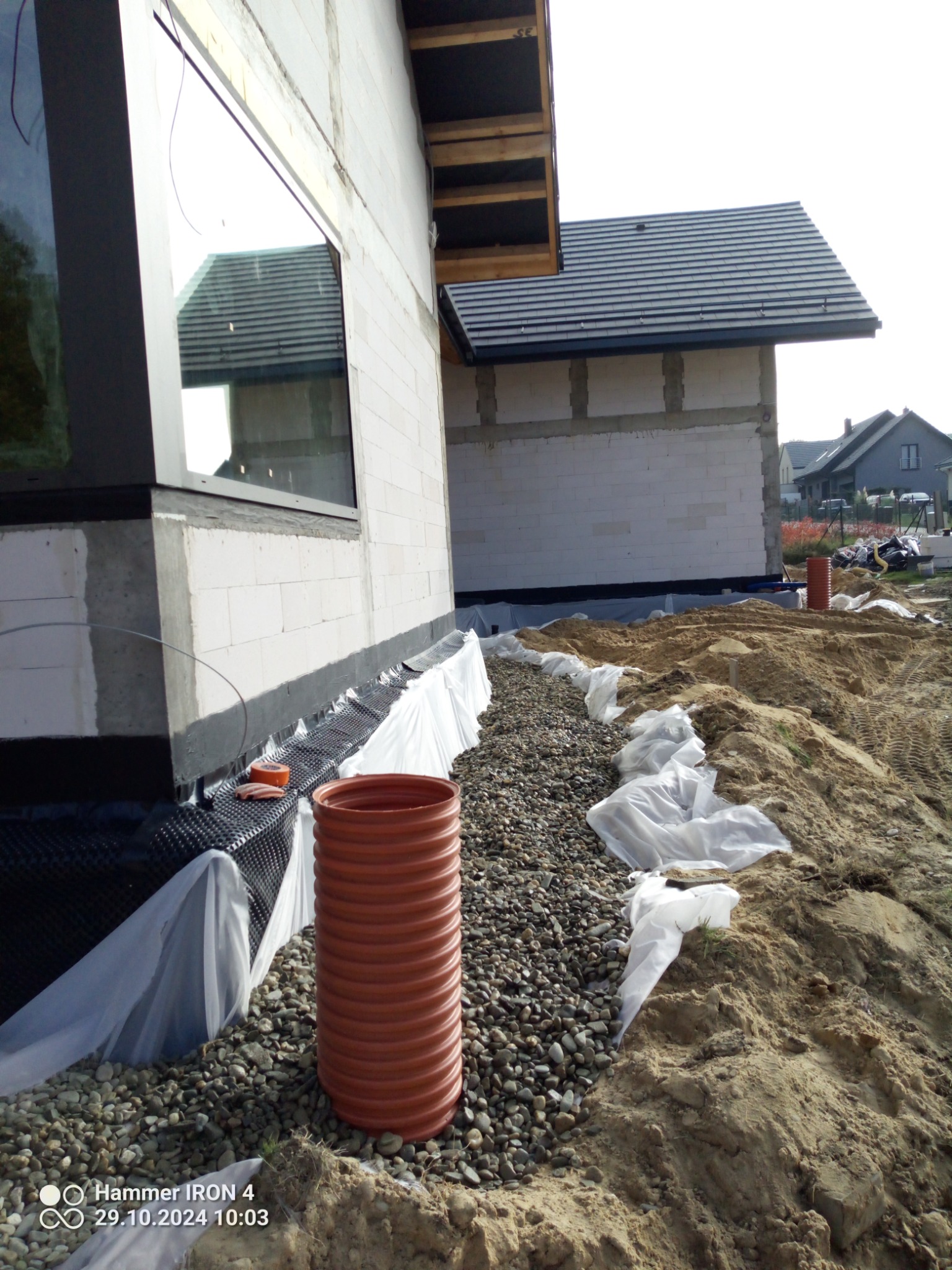 Fundamenty domu w trakcie budowy, widoczny drenaż opaskowy z rurą drenarską, folią i warstwą żwiru, ściany z pustaków oraz okno z odbiciem dachu.