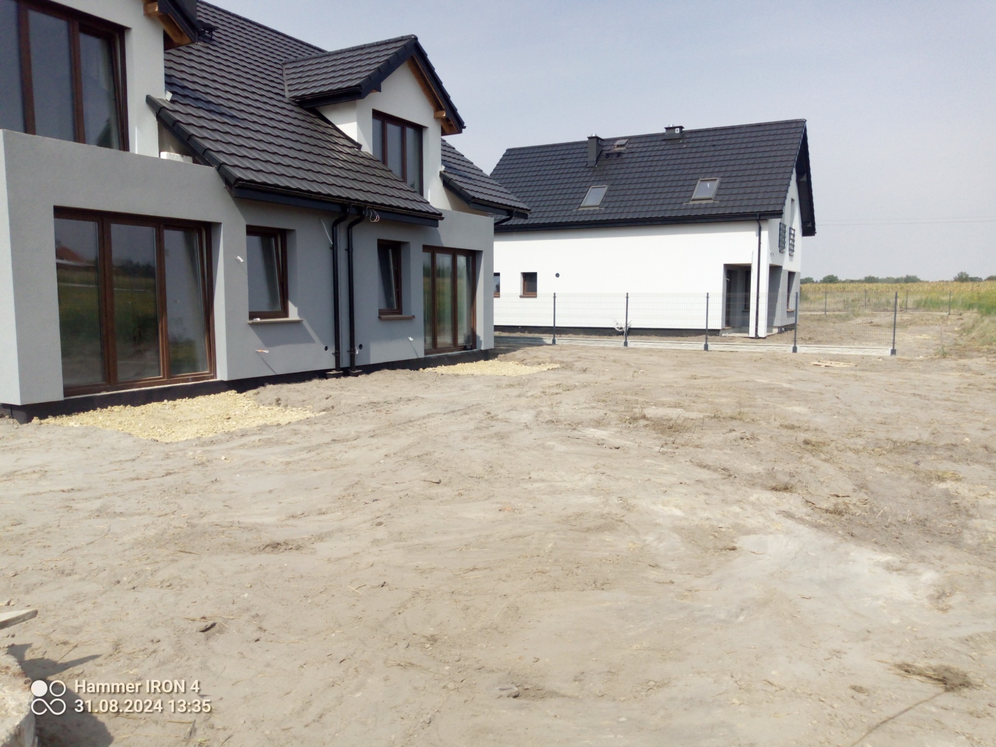 Dwa nowoczesne domy jednorodzinne w stanie surowym zamkniętym, z widocznym terenem przygotowanym pod zagospodarowanie, częściowo wysypanym żwirem.