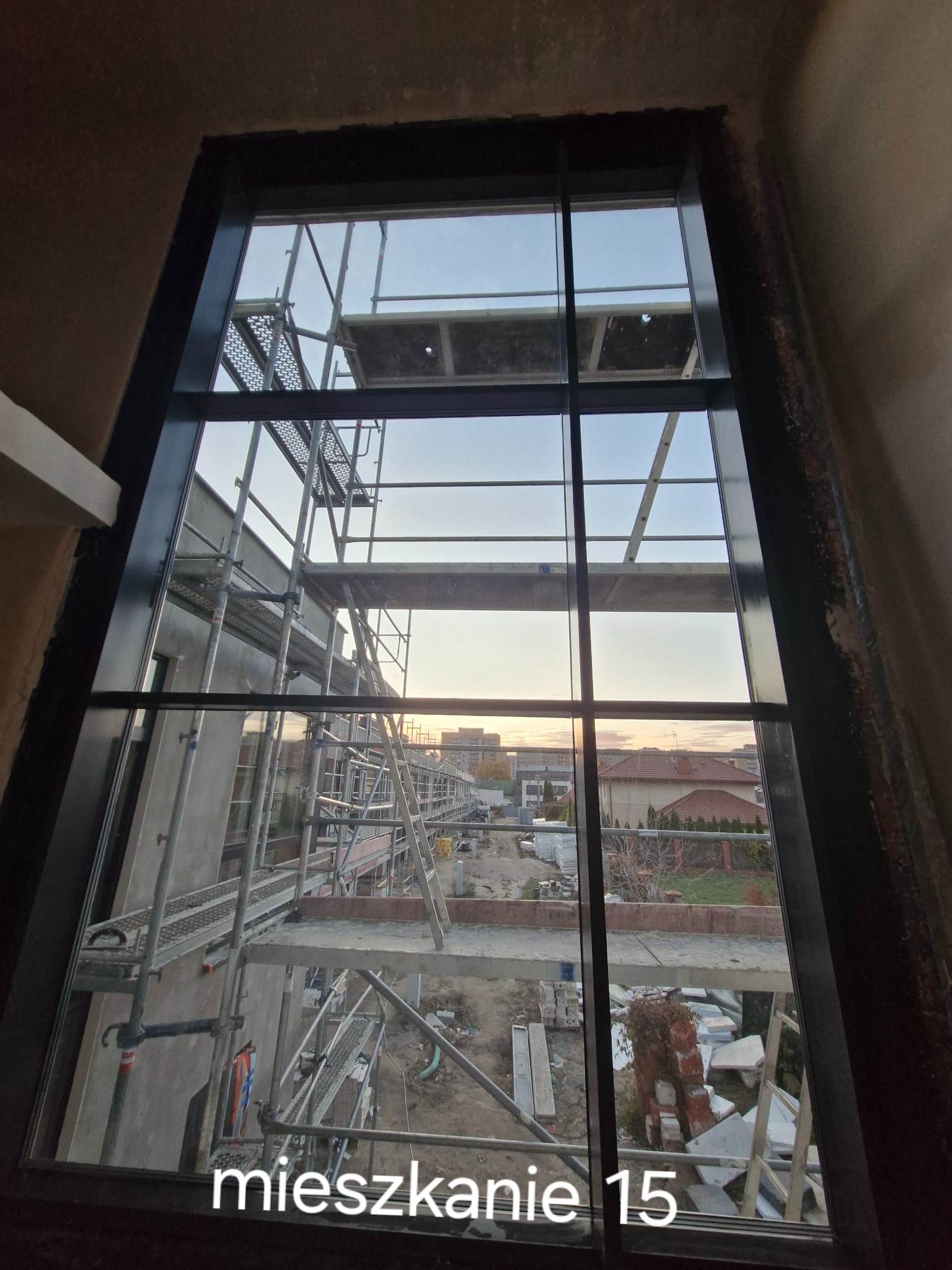 Wysokie okno z czarną ramą, widok na rusztowania i budynek w trakcie budowy, fragmenty gruzu na zewnątrz, wieczorny krajobraz w tle.