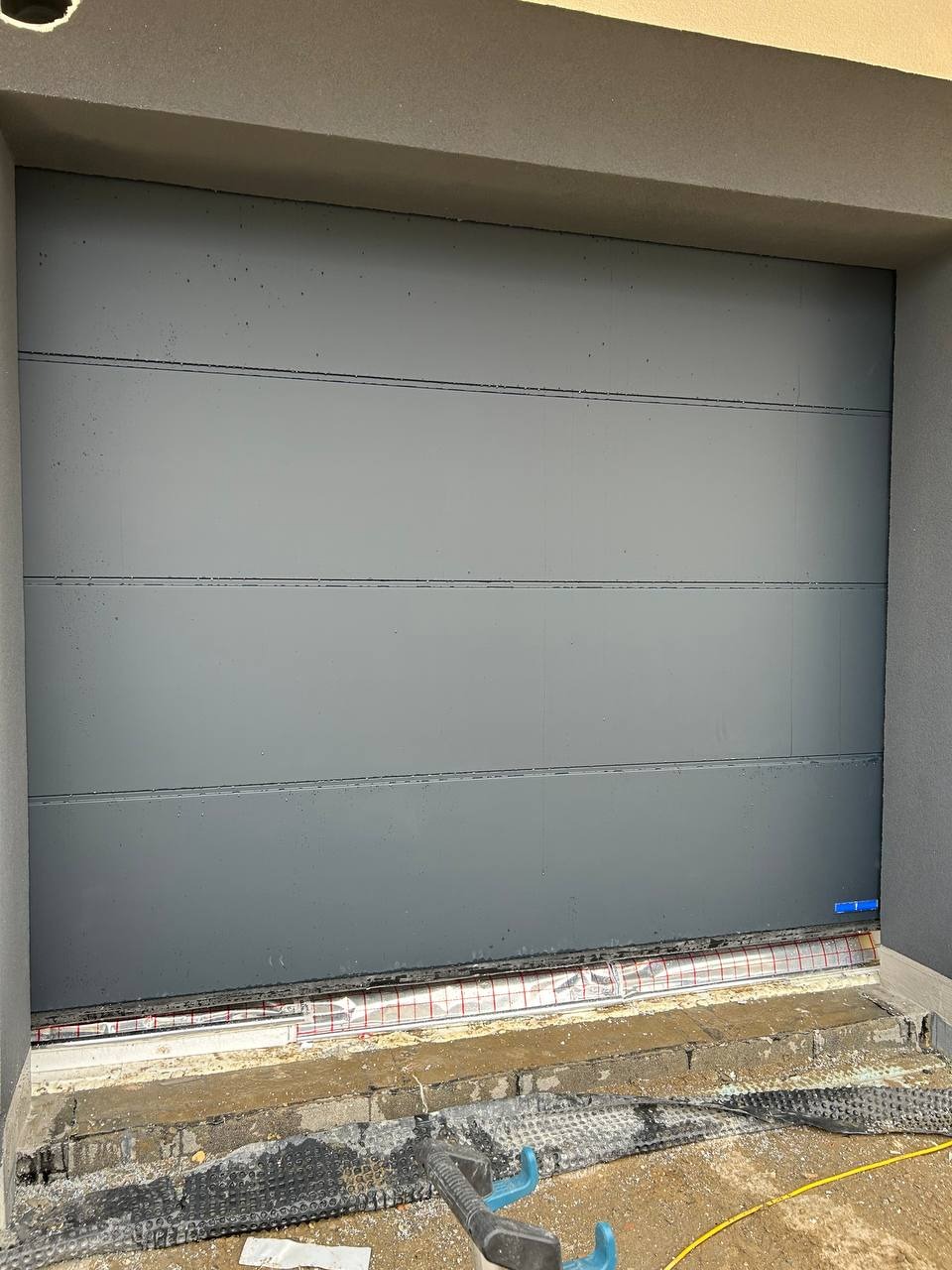 Szara, segmentowa brama garażowa w trakcie montażu, widoczne elementy izolacyjne i narzędzia na posadzce.