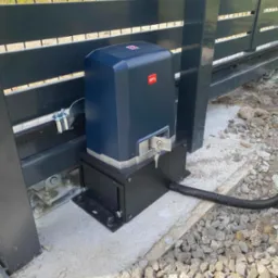 Zamontowany napęd bramy przesuwnej BFT z kluczem w zamku, widoczny przewód zasilający i fragment bramy z metalowymi profilami.